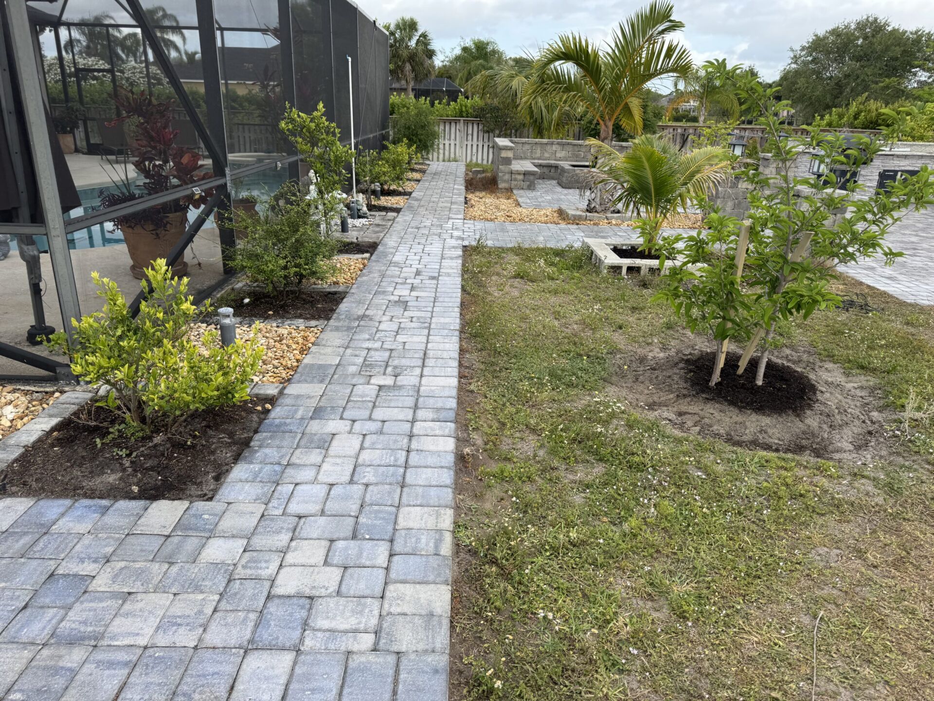 Gray paver path beside planted beds and a screened pool enclosure.