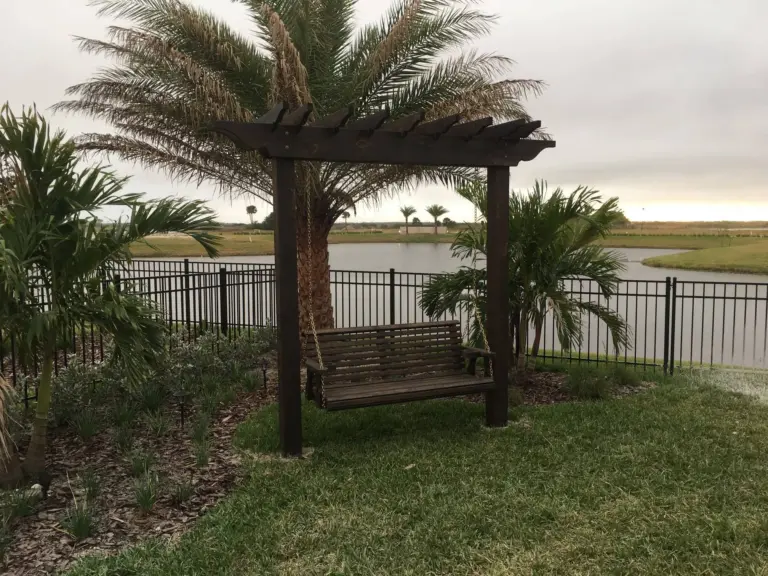 Wooden swing beneath an arbor beside palms and a fenced pond.