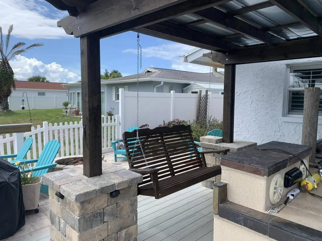 Dark wooden outdoor swing bench suspended beneath a covered pergola beside a home.