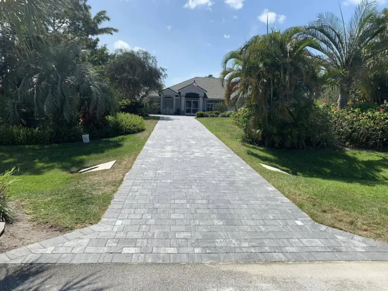 Long gray paver driveway bordered by lawn and palms leading toward a home.