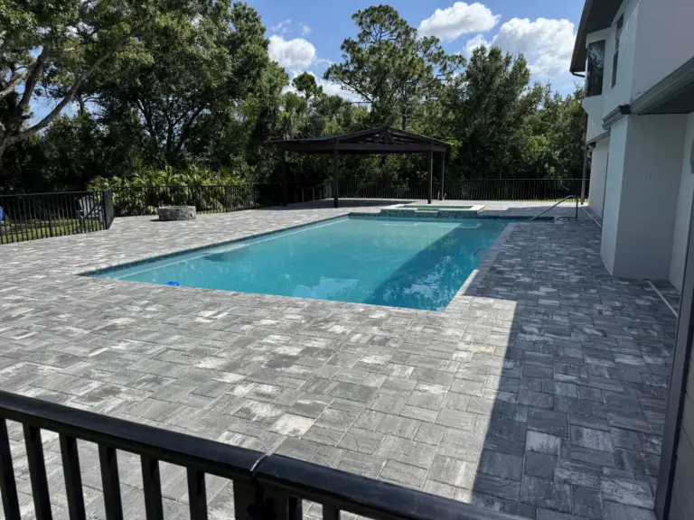 Wide gray paver deck surrounding a rectangular pool beside a white home.
