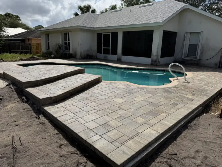 Gray pool deck pavers with dark borders and broad steps beside a curved backyard pool.