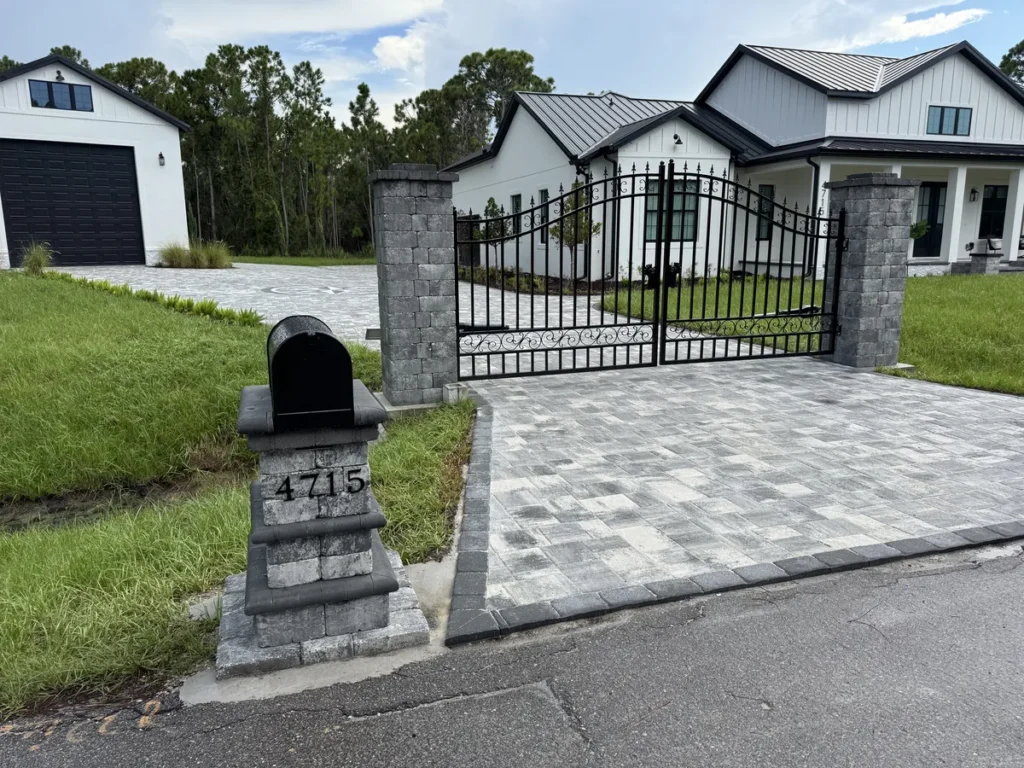 Gray paver driveway with a charcoal border at a gated entrance beside a white home
